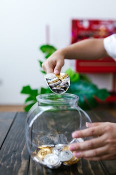 Hands placing bitcoins into a glass jar on a wooden table, symbolizing savings.