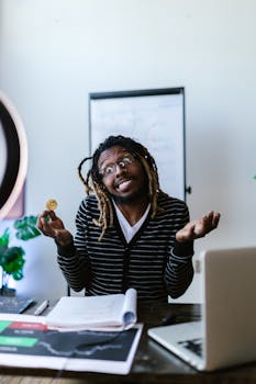 A man with dreadlocks and eyeglasses poses with a Bitcoin coin, expressing uncertainty in a financial setting.