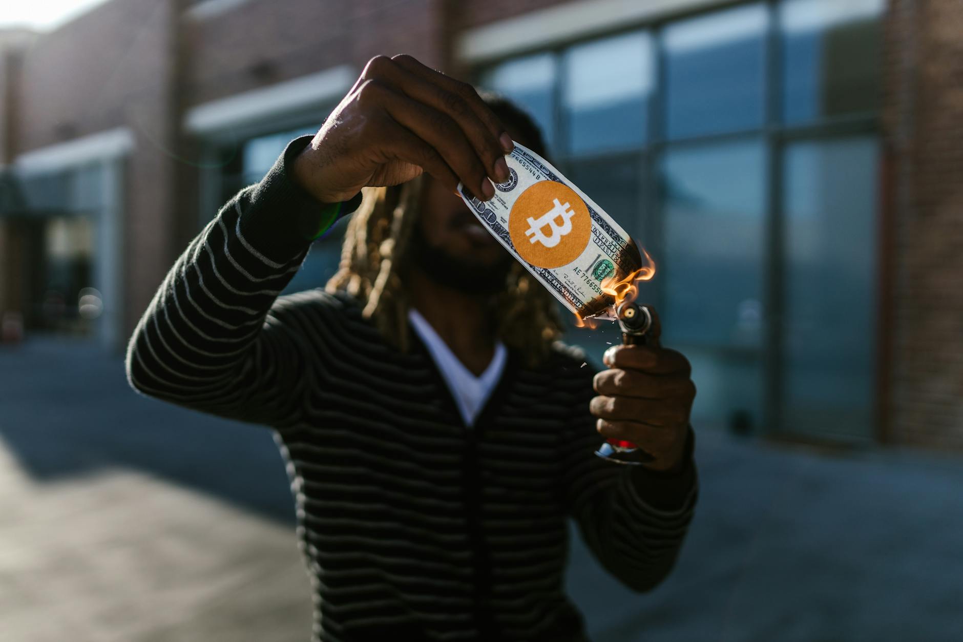 A man burns a fake dollar with a Bitcoin symbol outdoors, symbolizing cryptocurrency risks.