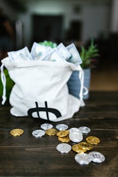 A bag overflowing with currency and scattered coins on a wooden table, symbolizing wealth.