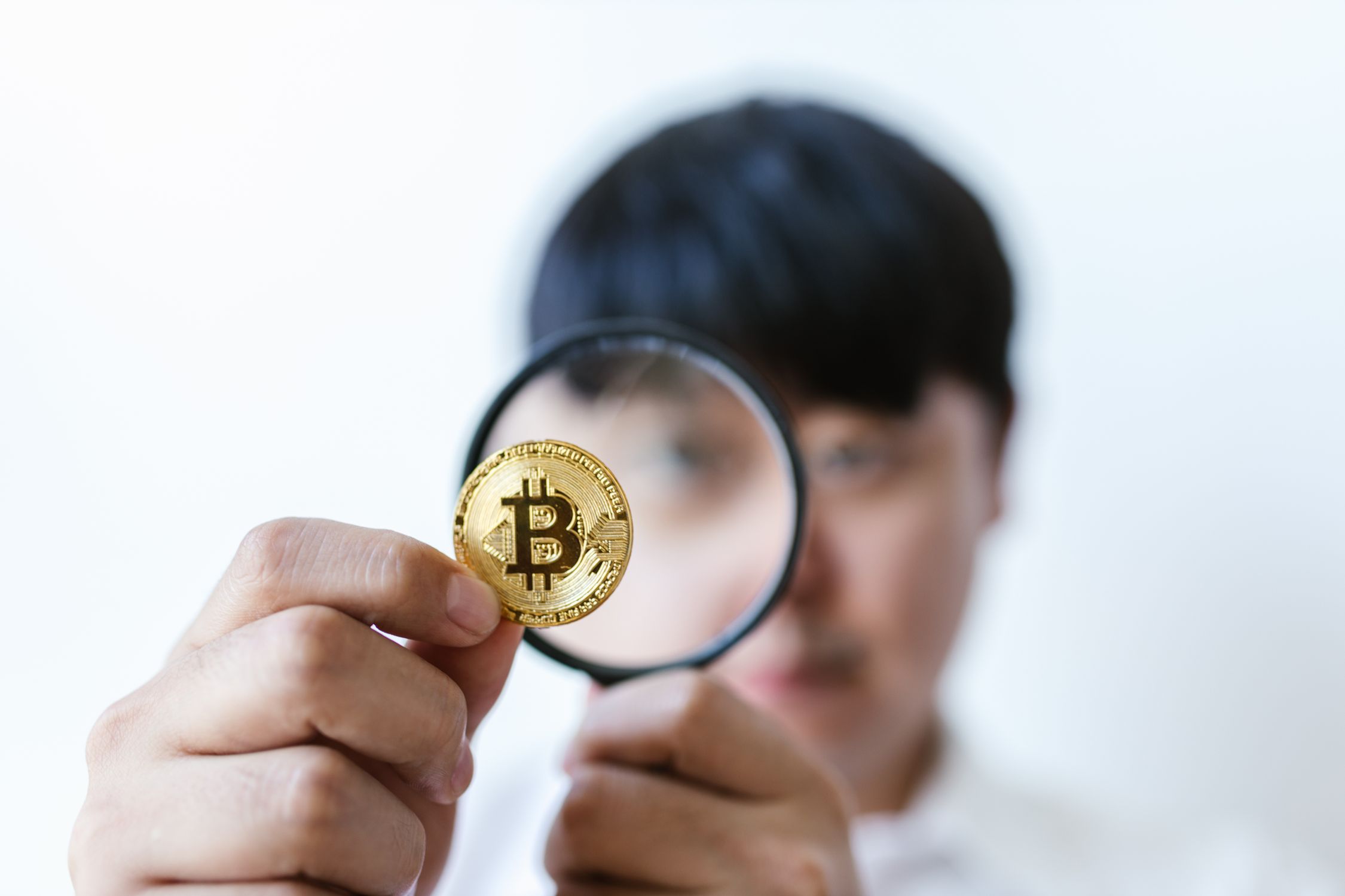 Pessoa examinando Bitcoin com lupa, simbolizando a análise dos mecanismos de consenso PoW e PoS.