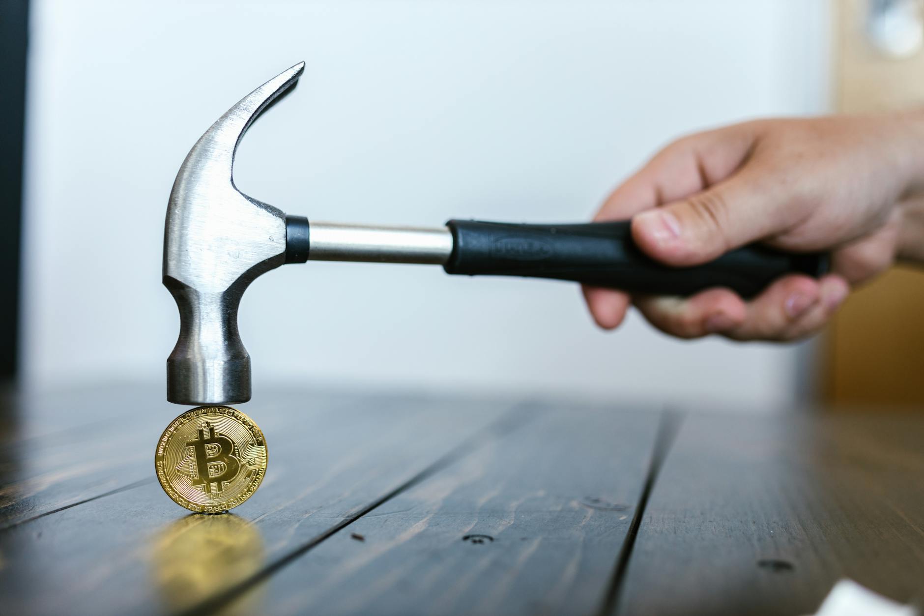 A hand holding a hammer above a Bitcoin coin on a wooden surface, symbolizing cryptocurrency volatility.