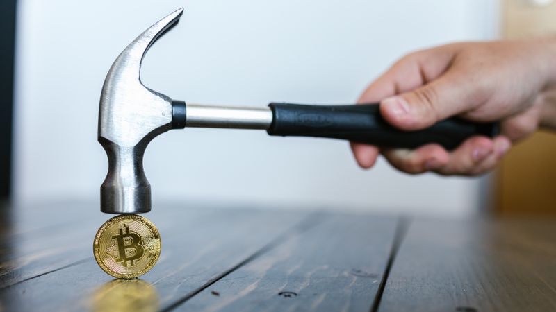 A hand holding a hammer above a Bitcoin coin on a wooden surface, symbolizing cryptocurrency volatility.