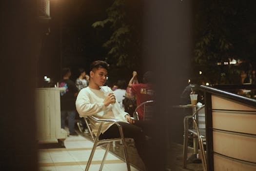 A young man enjoying a drink at an outdoor cafe at night, sitting on a terrace.