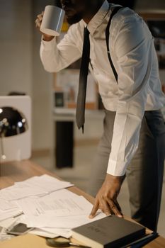 Businessman in suit with suspenders drinking coffee while reviewing documents in office setting.