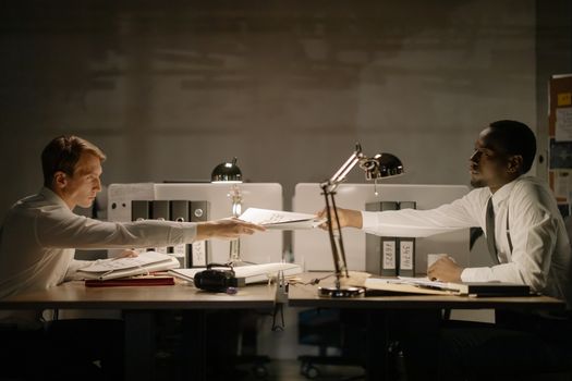 Two men in a professional office exchange documents under desk lamps, creating a focused work atmosphere.