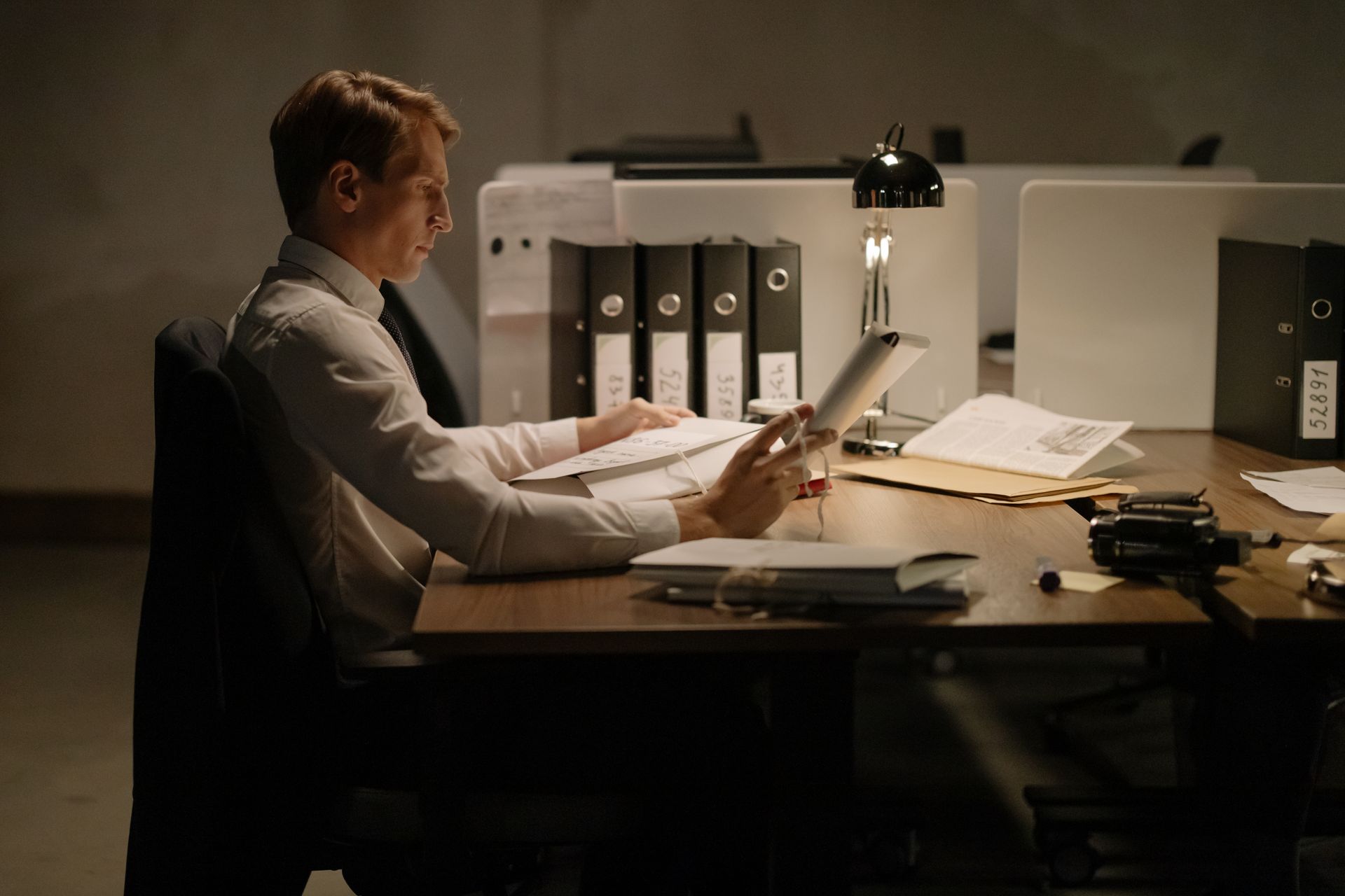 An office worker in formal attire intensely focused while reading documents at a desk with folders and a lamp.