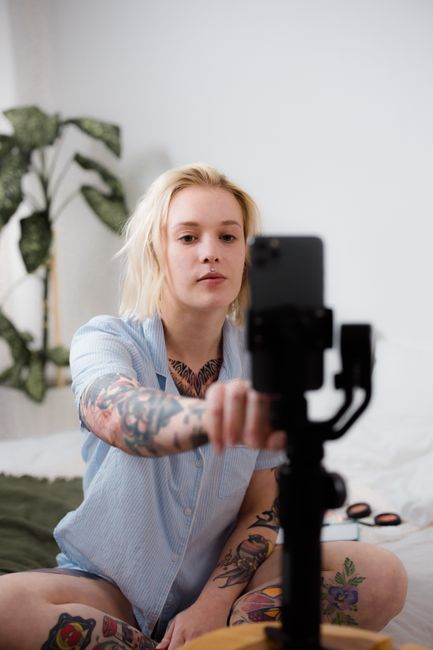 Tattooed woman with blonde hair taking a selfie indoors with a smartphone on a tripod.