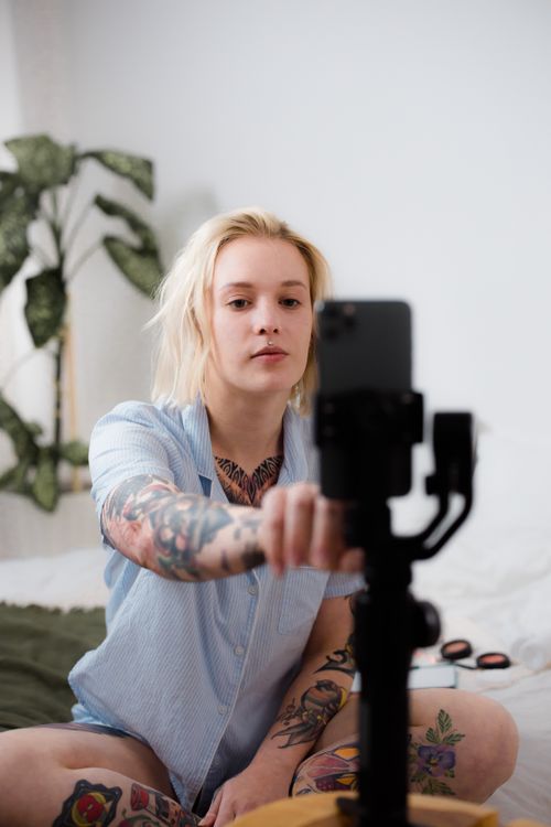 Free Tattooed woman with blonde hair taking a selfie indoors with a smartphone on a tripod. Stock Photo