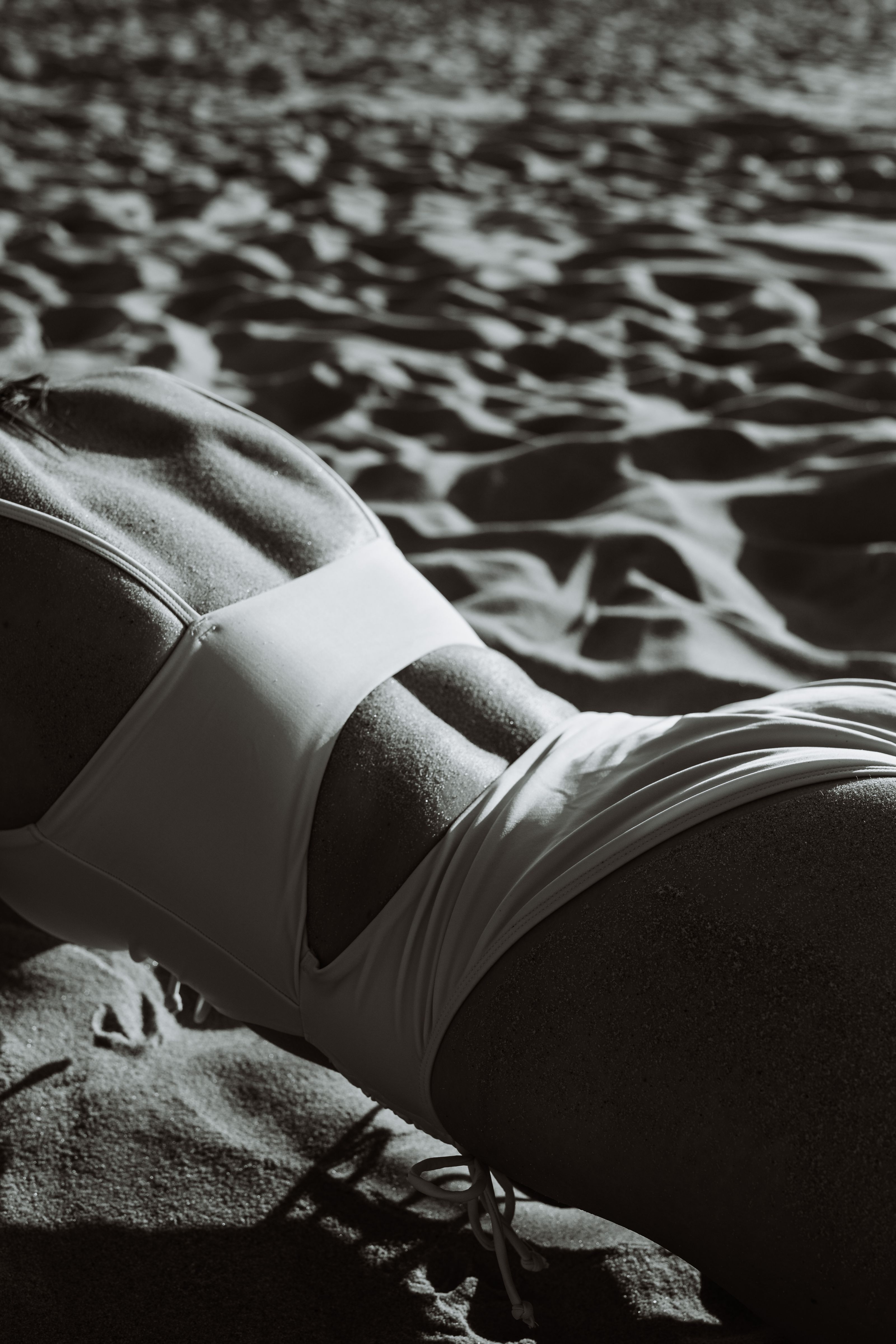 Uma imagem em tons de cinza de uma mulher de maiô deitada em uma praia de areia, enfatizando textura e contraste.