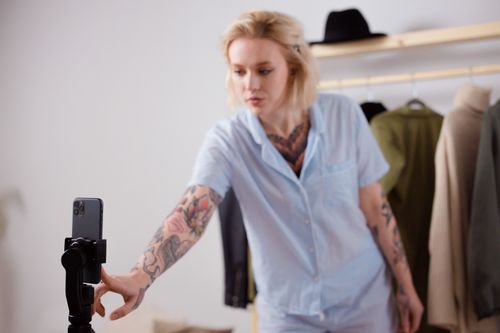Free A stylish woman with tattoos adjusting her smartphone on a tripod, surrounded by clothing. Stock Photo