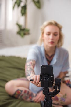 A tattooed woman uses a smartphone mounted on a gimbal indoors, depicting content creation.