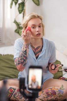 Tattooed woman applying makeup while filming tutorial in a cozy room.