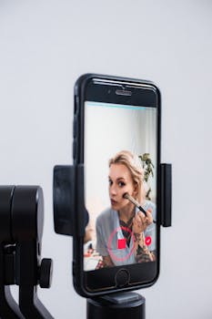 Blonde woman applying makeup, filming tutorial on smartphone using a phone holder.