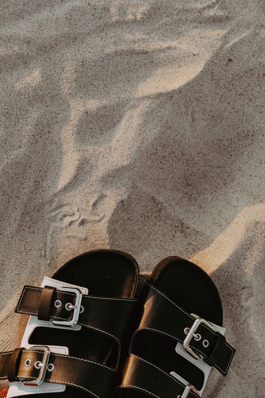 Black leather sandals resting on sand, echoing the way texture can make a minimal summer shoe look considered rather than precious.