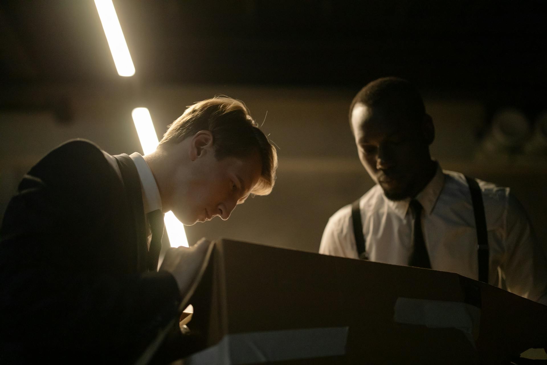 Two detectives examine a box in dimly lit room, focused on investigation.