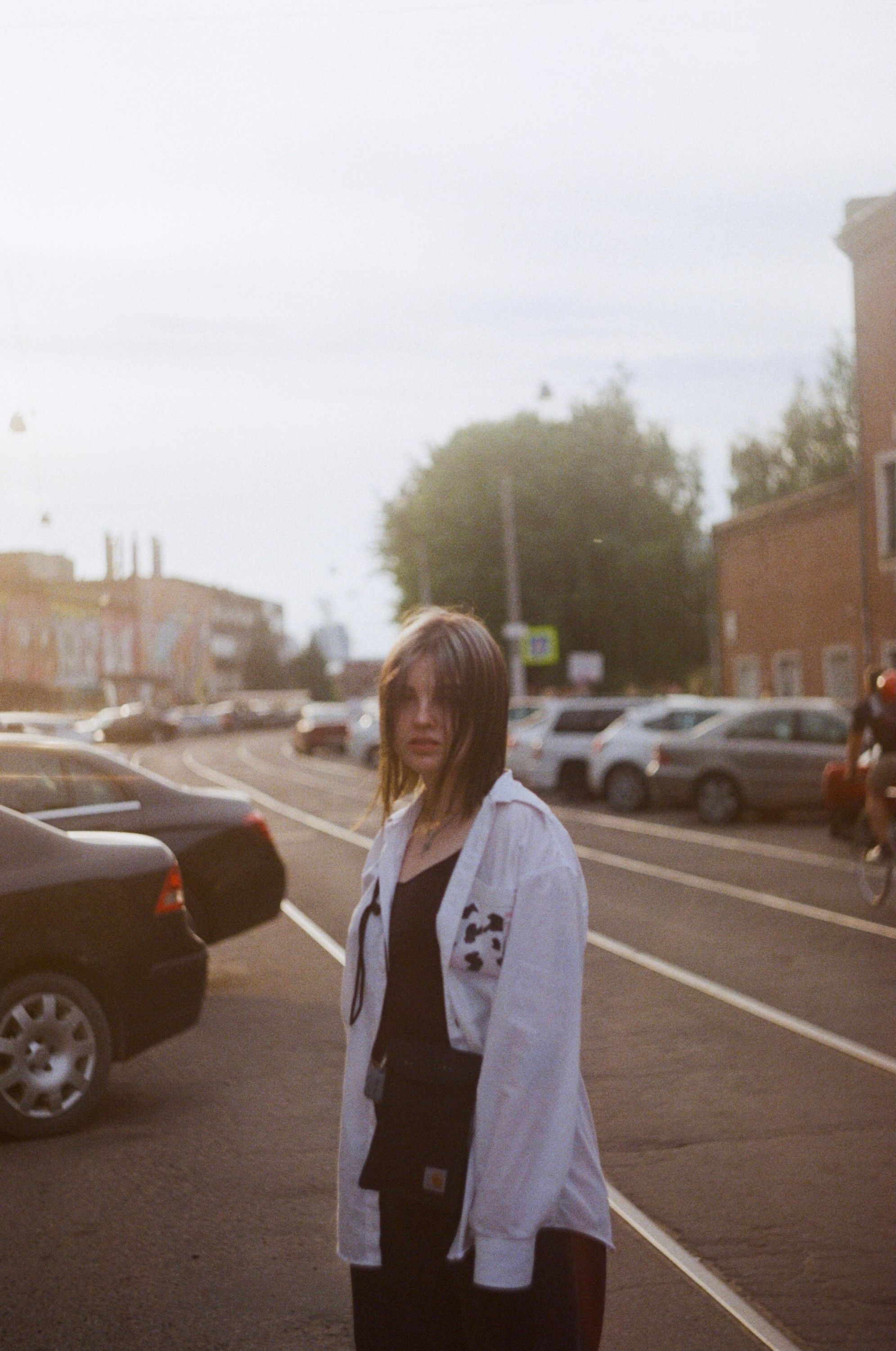 A young woman in a white shirt stands on a bustling city street with parked cars and tram tracks.