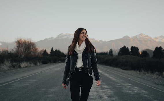A woman in a leather jacket joyfully walks along a scenic mountain road during the day.