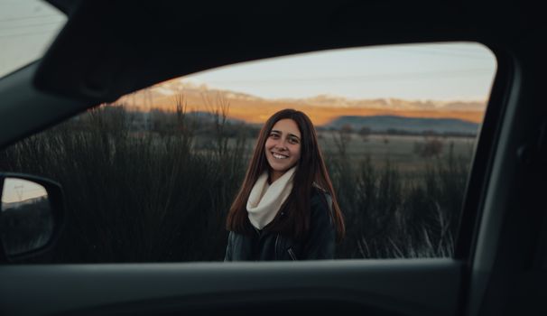 A woman smiles warmly while framed by a car window at sunset, capturing a serene moment.