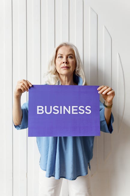 Confident senior woman presenting a business sign in a bright studio setting, wearing a casual blue outfit.