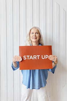 Elderly woman in blue shirt holding a 'Start Up' sign, promoting entrepreneurship.