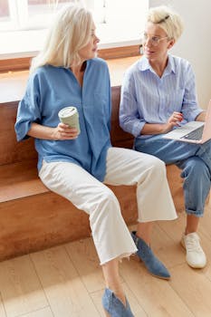 Two women relaxing indoors, sitting on a bench, enjoying a conversation with a laptop and tumbler.