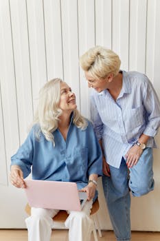 Two senior women sharing a joyful moment while using a laptop together indoors.