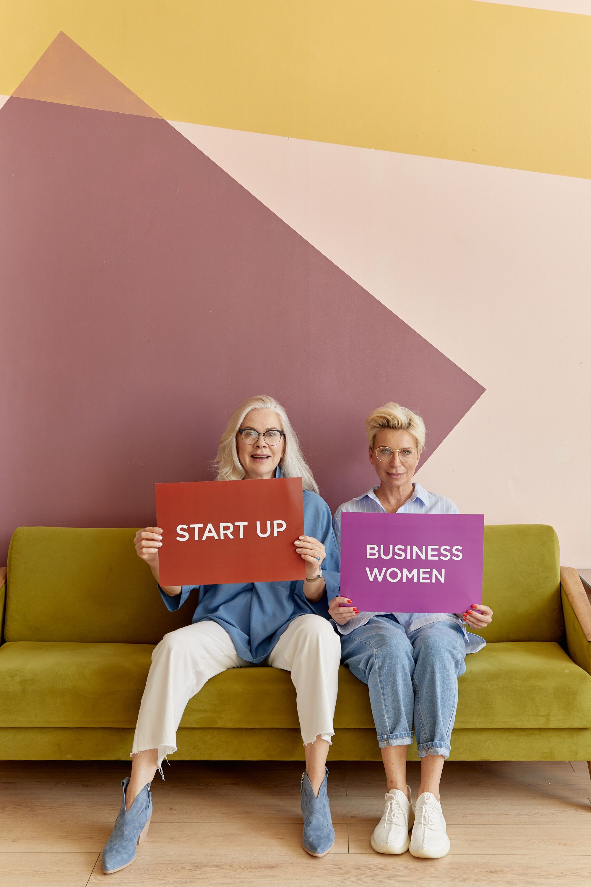 Two senior women holding startup and businesswomen signs, sitting on a sofa.