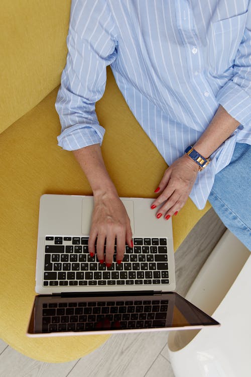 Person in Blue and White Striped Dress Shirt and Blue Denim Jeans Sitting on Sofa Using Laptop