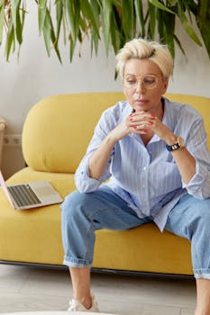 Thoughtful woman with eyeglasses sitting indoors on a yellow sofa, laptop beside her.