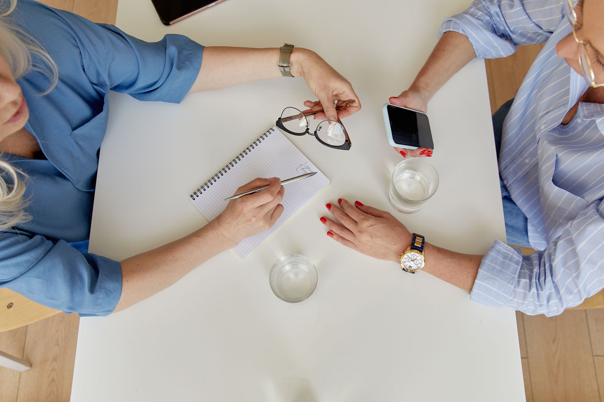 Two women in a business meeting taking notes with glasses and smartphone on a white table.