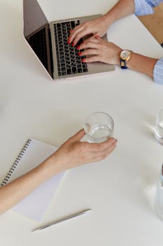 Overhead view of a modern workspace with a laptop, hands typing, and a glass of water.