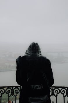 A solitary figure in a raincoat overlooks a foggy Istanbul from a balcony.