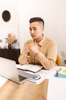 Focused Asian businessman working at desk with laptop and documents in a modern office setting.