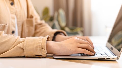 Close-up of hands typing on a laptop indoors, wearing a casual jacket.