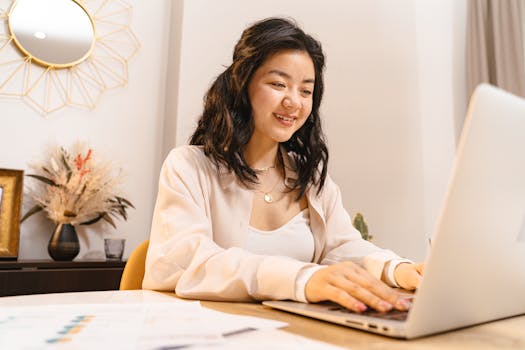 Smiling Asian woman typing on a laptop at home office with decor and papers.