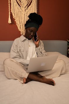 African woman sitting on bed using laptop and phone, working from home. Cozy indoor setting.