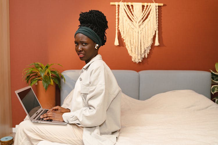 A Woman In White Denim Jacket Sitting On The Bed While Using Her Laptop