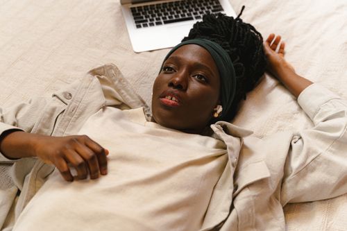 Free A woman with long hair rests on a bed beside a laptop, appearing contemplative and relaxed. Stock Photo