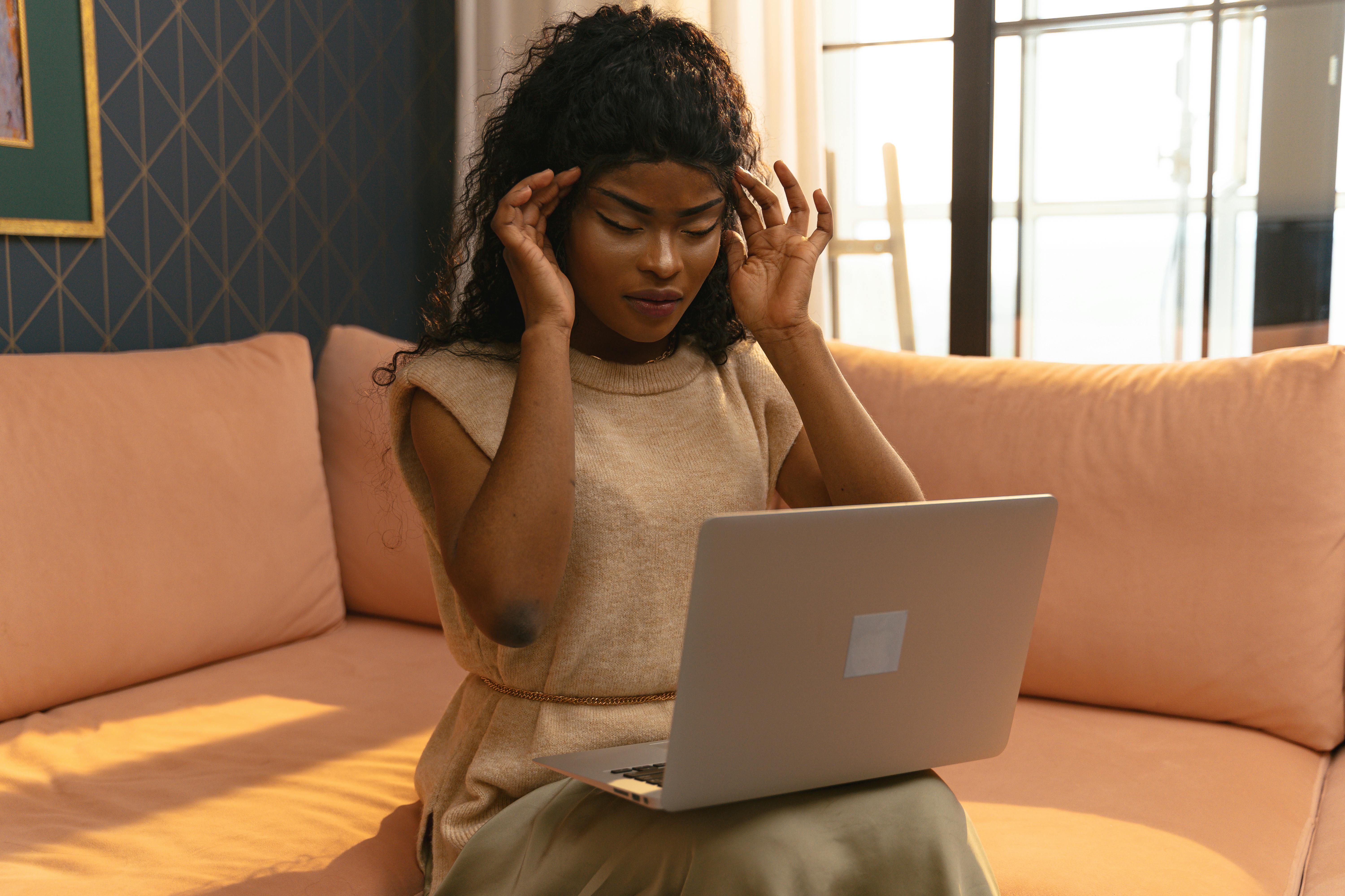 A young woman holding her head in pain while working on a laptop in a cozy home interior.