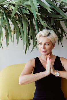 A blond woman sits indoors practicing yoga with a serene expression.