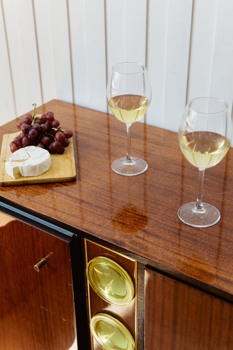 Glasses Of White Wine On Brown Wooden Cabinet Beside Grapes And Cheese On Wooden Board