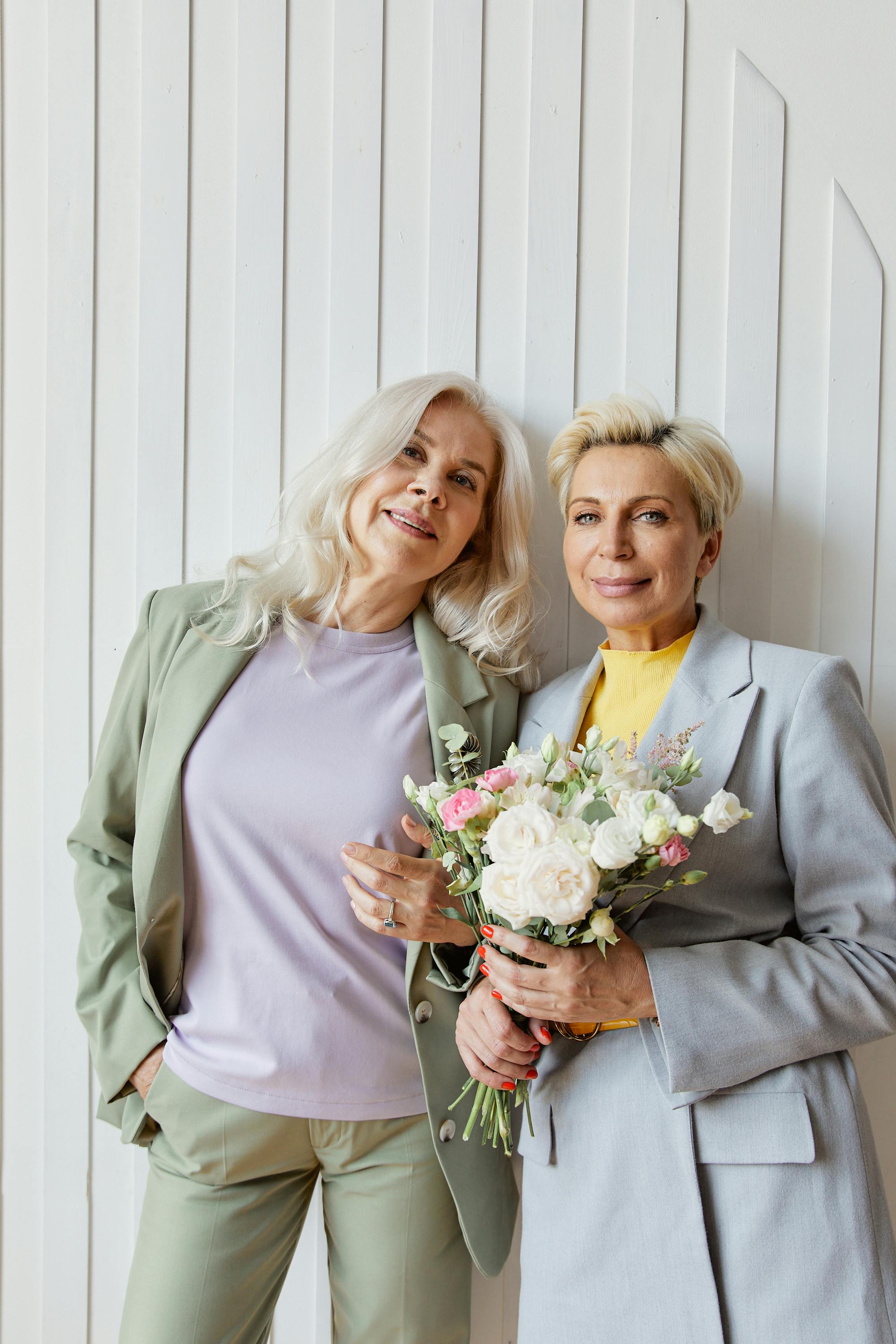 Two stylish senior women posing with a flower bouquet against a textured white wall.