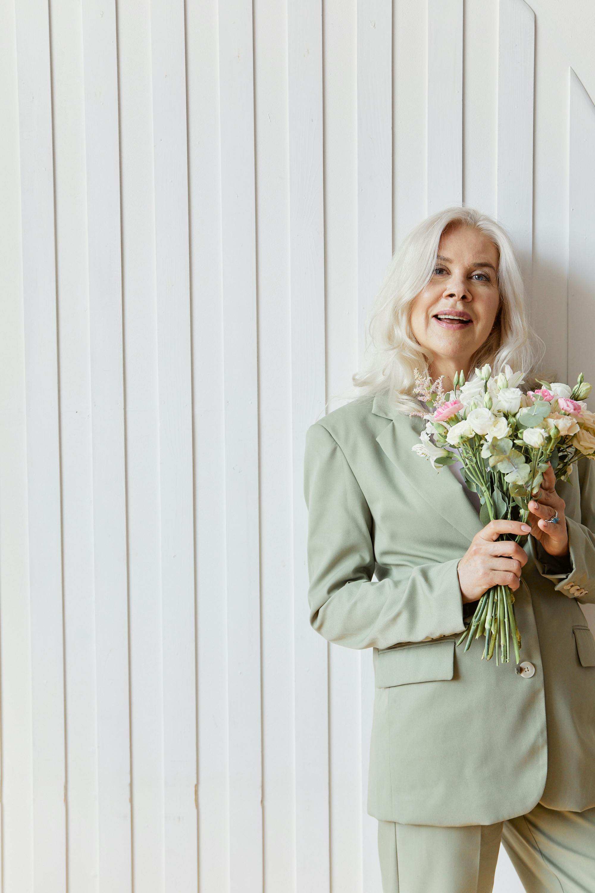 A stylish senior woman with white hair in a suit holding a bouquet of flowers indoors.
