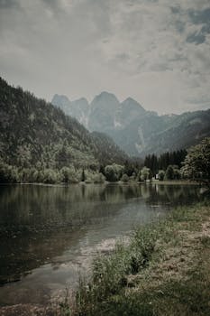 A tranquil mountain lake surrounded by lush greenery under a moody, overcast sky.