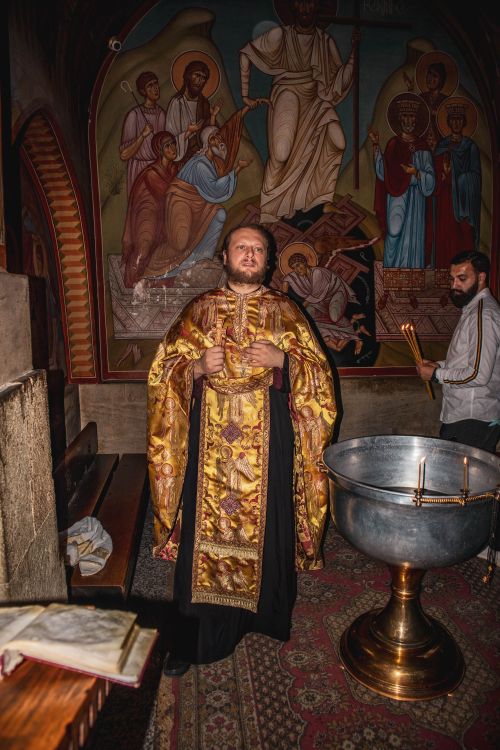 Free Orthodox priest in ornate robe stands in front of religious mural preparing for baptism. Stock Photo