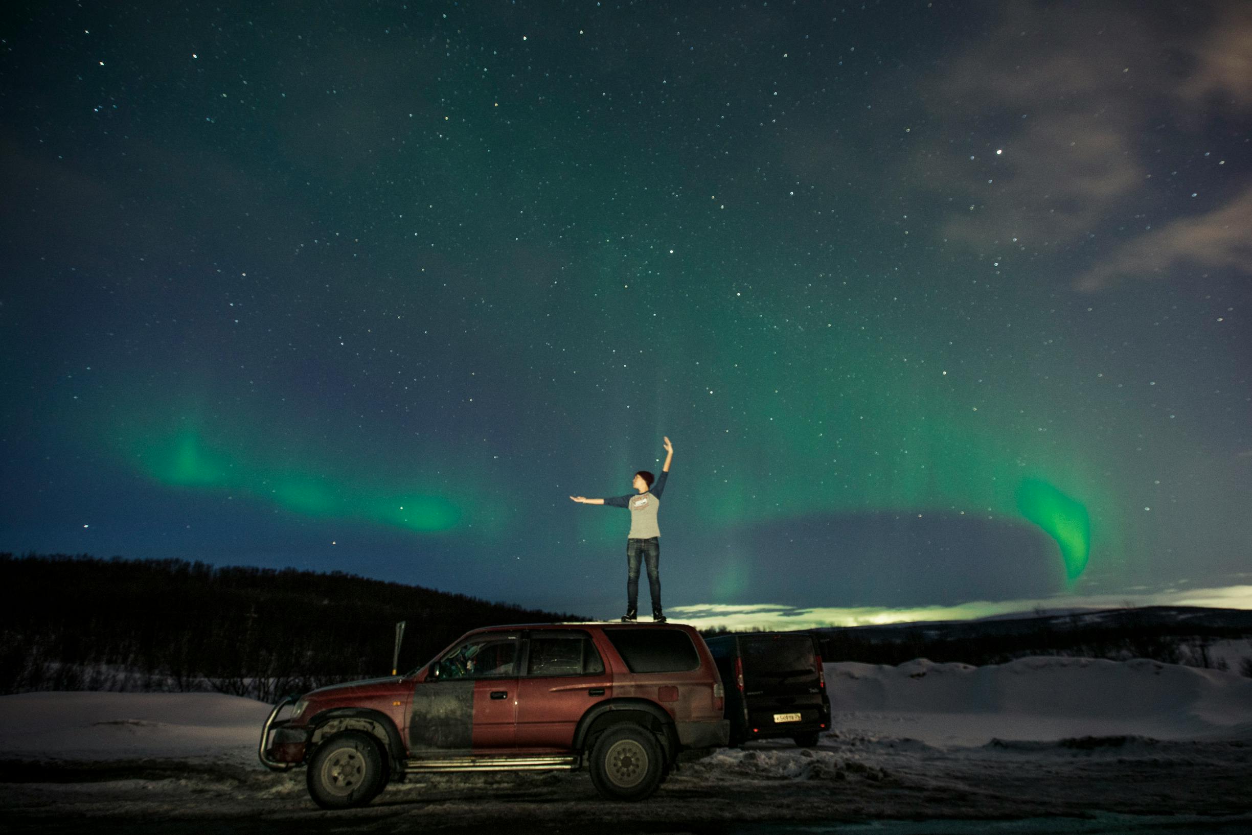 Person stands on SUV under Northern Lights, embracing winter night adventure.