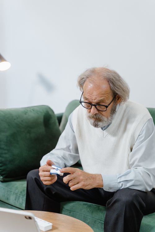 Free Elderly man in glasses sits on a green sofa holding an object, indoors. Stock Photo
