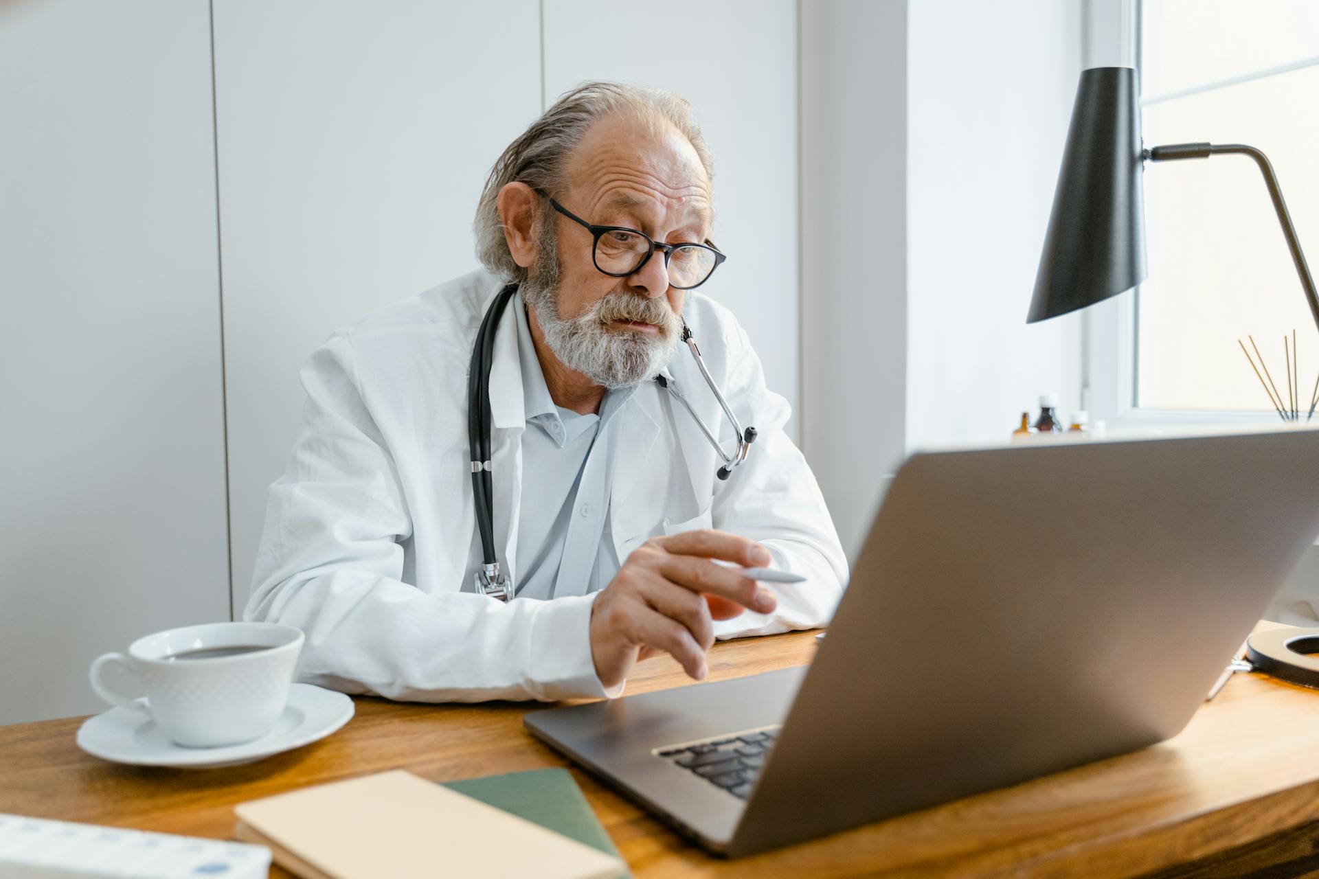 Senior male doctor using laptop for telehealth consultation in office setting.