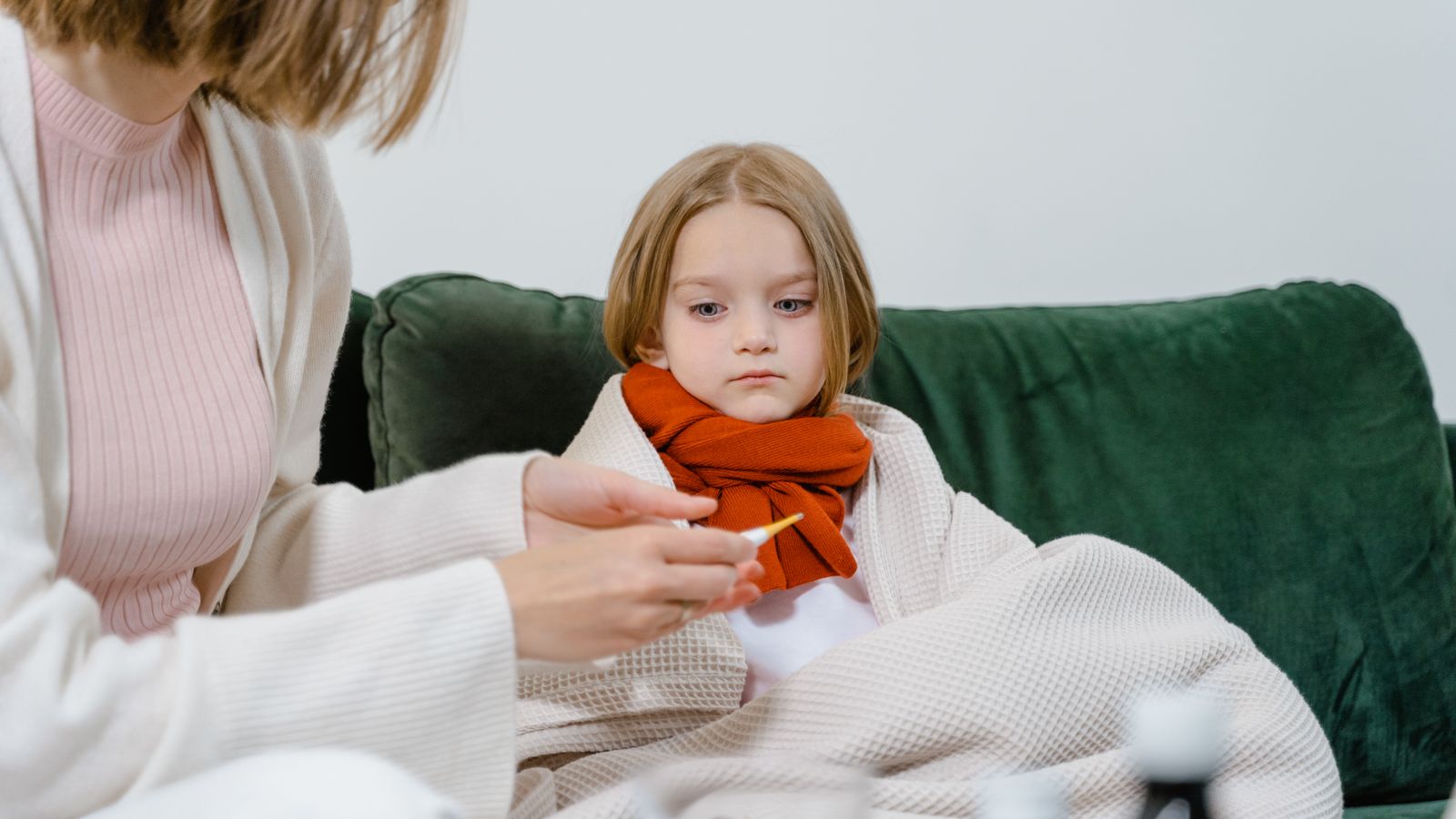 A caring moment of a woman checking a sick child's temperature on a cozy green couch indoors.